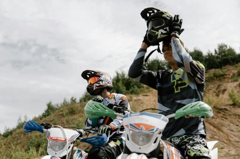 Two riders on dirt bikes preparing for an off-road motocross adventure under a cloudy sky.